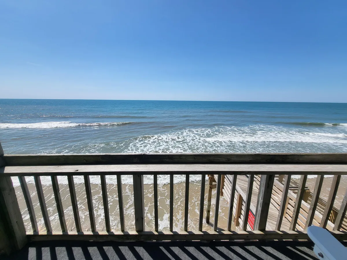 A serene view from the balcony shows ocean waves, a wooden railing, and sunlight streaming across the water. The peaceful mood evokes the experience of relaxing outdoors, listening to the waves, and enjoying sunsets or stargazing.