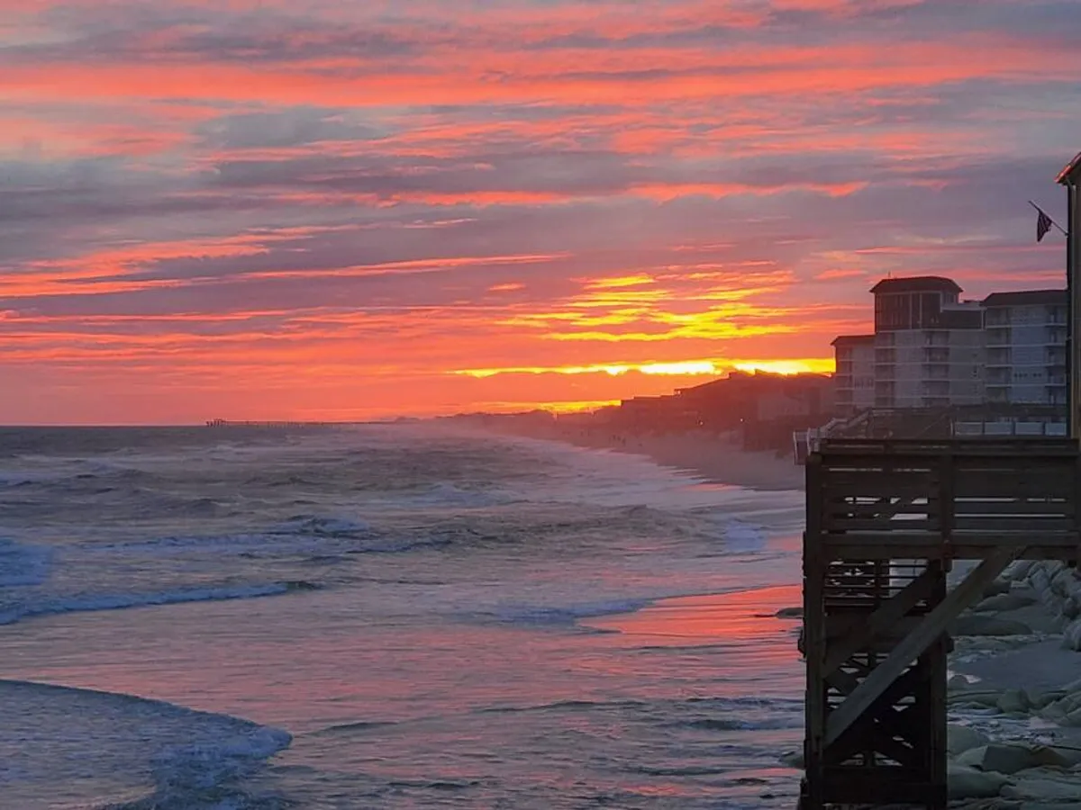A stunning view of the sun setting over the ocean, with a silhouette of a pier and buildings along the shoreline. The scene is tranquil and inviting, perfect for capturing the magical atmosphere of an evening beach walk.