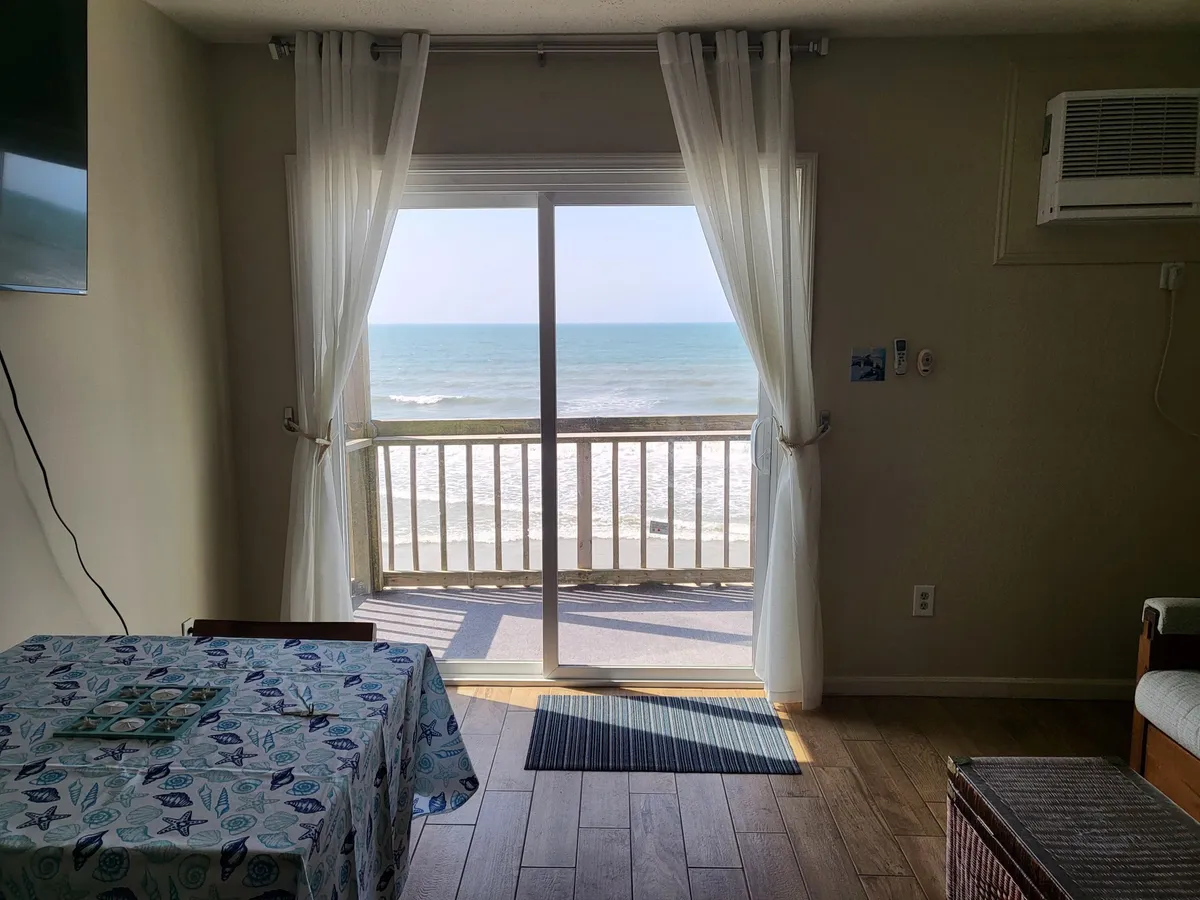 The image presents an open living and dining area with a large sliding glass door opening to expansive ocean views. The seating area is arranged to maximize enjoyment of the scenery, and the dining table is perfectly positioned for meals with a view.