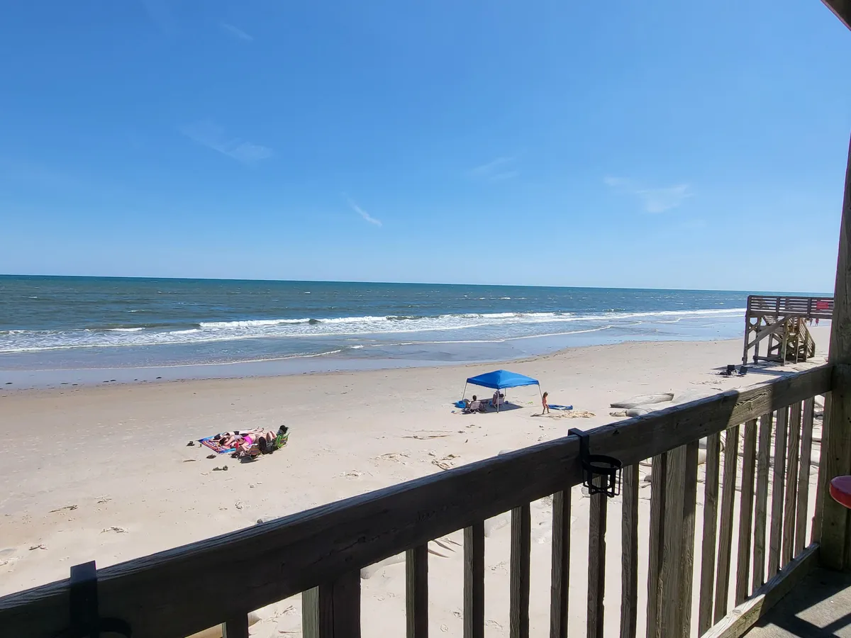 A bright, inviting scene of the beachfront with golden sand, shimmering blue ocean, and a colorful beach umbrella under sunny skies. Families are seen enjoying a range of activities such as sunbathing, swimming, beach games, and picnicking, while children build sandcastles and search for seashells.