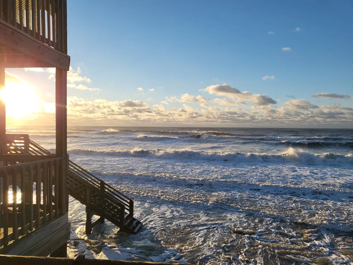 This image captures a breathtaking sunset over the ocean, with waves gently crashing onto the shore and wooden steps leading directly to the beach. The sky is ablaze with warm colors, creating a serene and romantic atmosphere perfect for evening strolls.