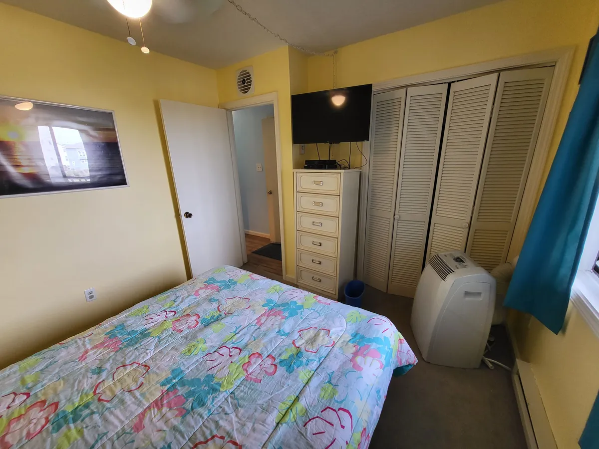 An inviting bedroom with a cheerful floral comforter on the bed, a dresser with ample storage, a wall-mounted TV, and a window adorned with blue curtains. The space exudes warmth and comfort, ideal for both relaxation and entertainment.