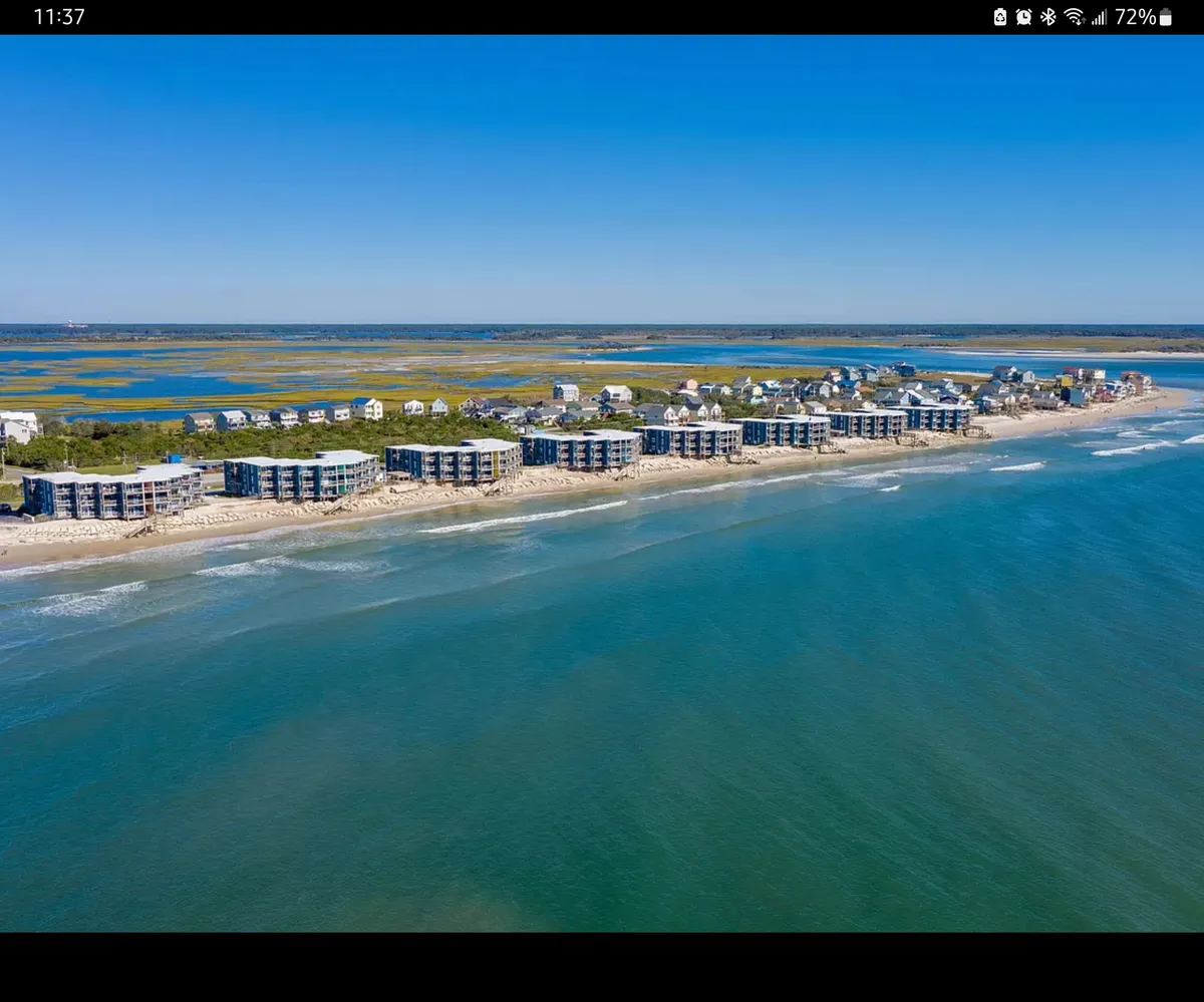 A wide-angle exterior shot of beachfront properties facing the ocean under a clear blue sky. The scene captures the relaxed vibe of the shoreline, with waves gently lapping and the promise of sunrise views from the property.