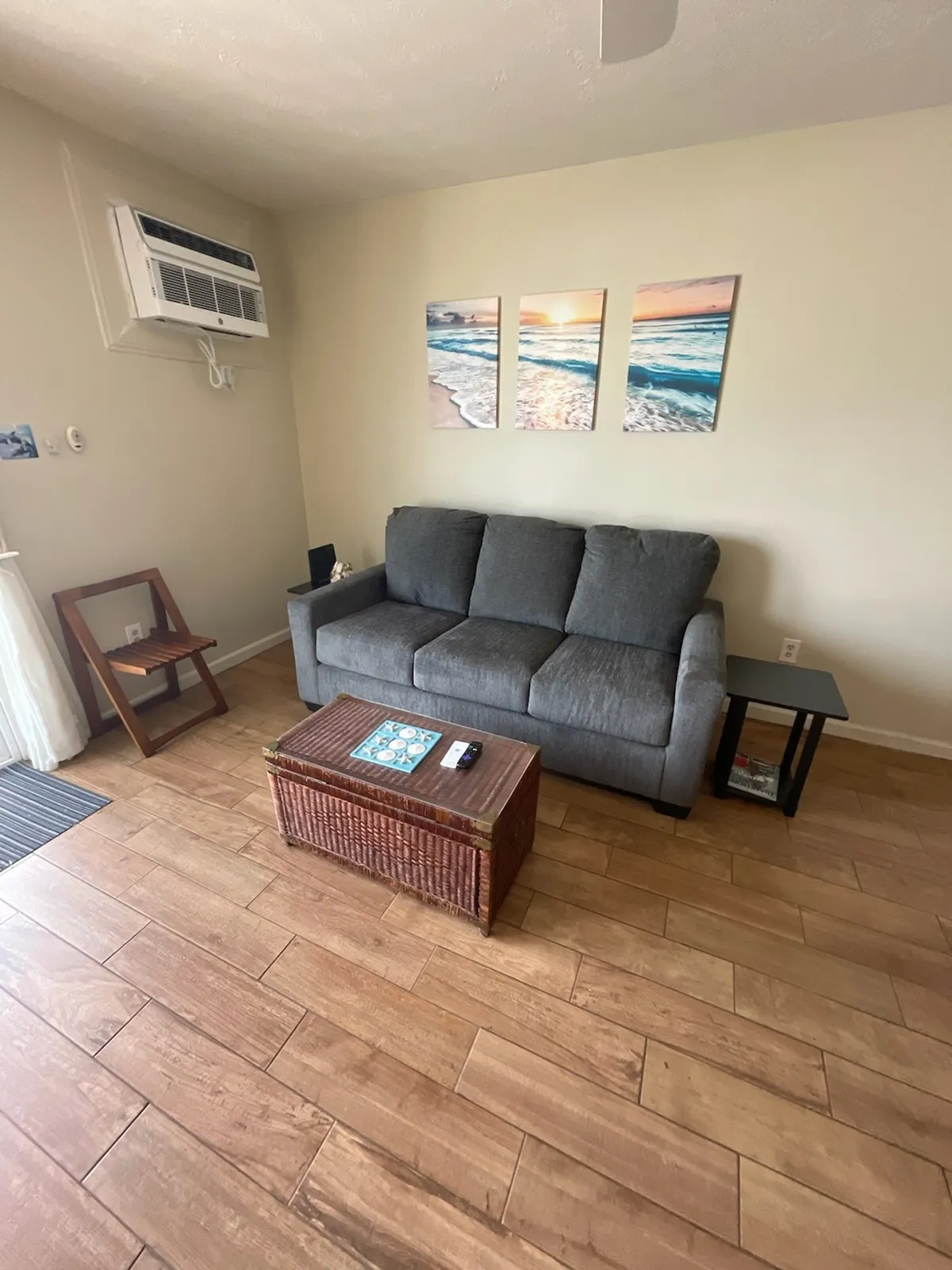 A cozy living room with a gray sofa, woven coffee table, ocean-themed wall art, and a small side table. The space feels bright and inviting, with touches of beach-inspired decor and plenty of natural light.