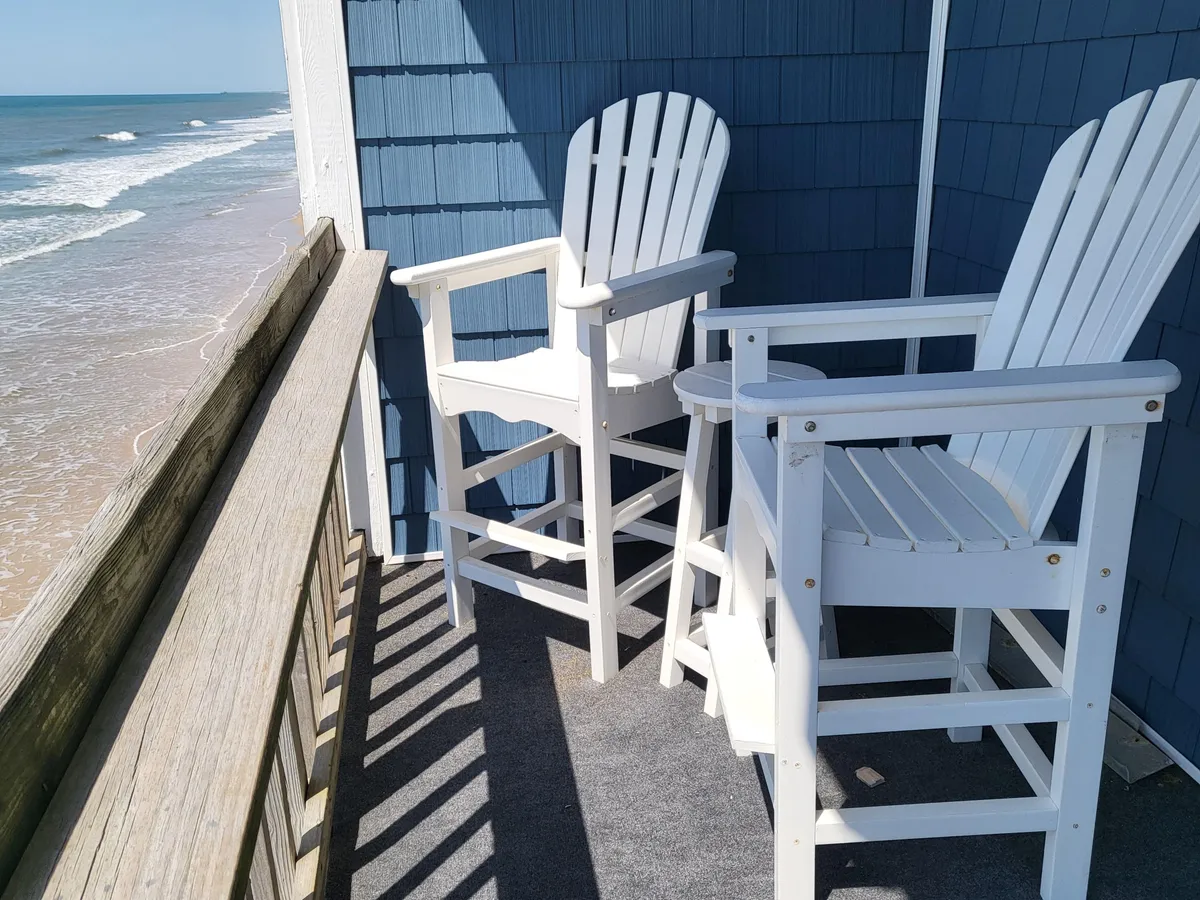 The hero image showcases a pair of inviting white Adirondack chairs on a private balcony, framed by a sturdy wooden railing and overlooking the endless blue expanse of the Atlantic Ocean. The tranquil scene suggests moments of calm, whether greeting the sunrise with a cup of coffee or unwinding as the stars appear, all while enjoying the soothing sound of the waves just steps away.