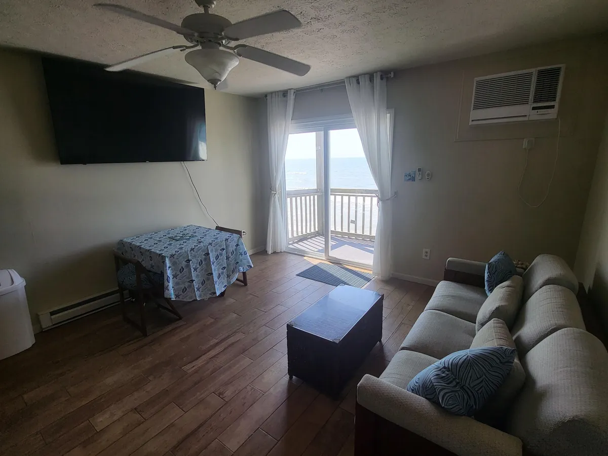 This image captures a comfortable living room with a large TV, a cozy sofa, a dining table, and a glass balcony door offering views out to the ocean. The space is bright and inviting, with natural light streaming in through the glass door and easy access to the balcony.