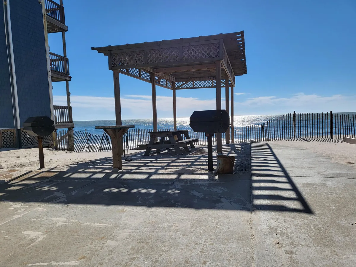 A welcoming outdoor area featuring a wooden gazebo, picnic table, and grills overlooking the ocean. The scene is ideal for sunset barbecues, casual meals, and gatherings with family or friends under the open sky.