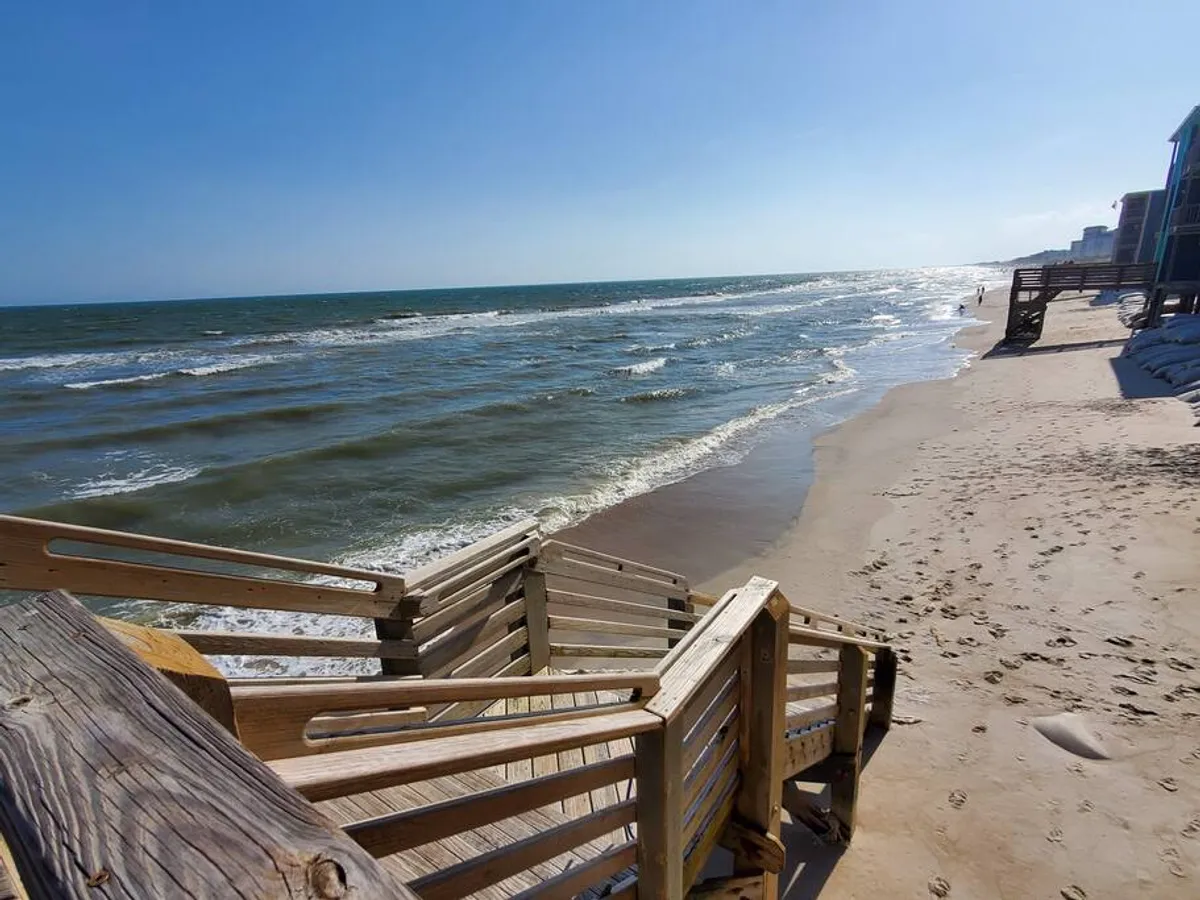 A tranquil scene featuring a wooden staircase leading directly onto the sandy beach, with gentle ocean waves rolling in. The shoreline stretches invitingly, ideal for morning walks or unwinding after work.